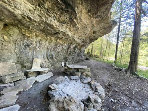 A rocky overhang with stone seating and a fire pit, surrounded by trees in a serene outdoor setting.