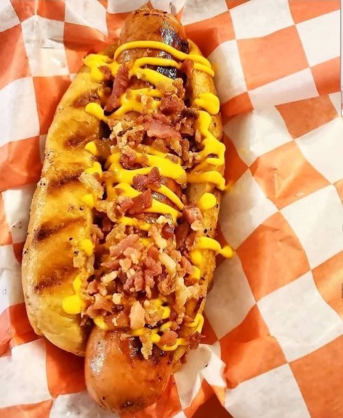 A grilled hot dog topped with chopped bacon and yellow mustard, served in a paper tray with orange and white checkered pattern.