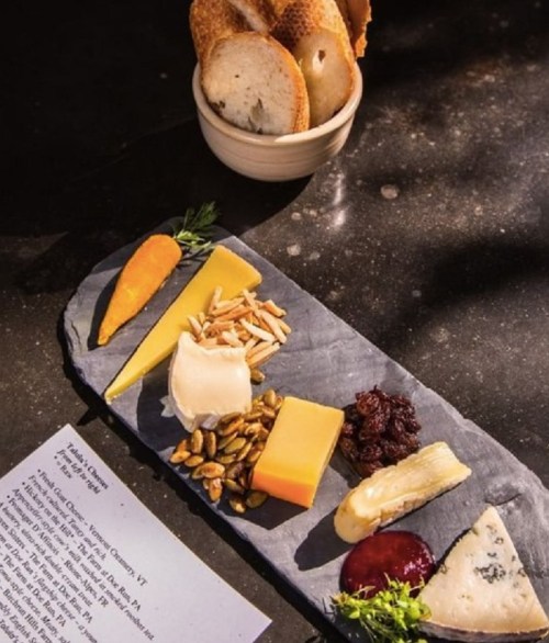 A slate cheese board featuring various cheeses, nuts, dried fruit, and a carrot, with a side of bread.