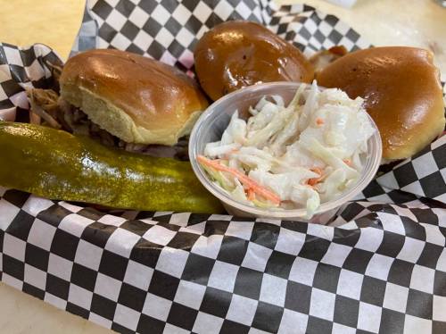 A basket with three pulled pork sliders, a pickle, and a side of coleslaw on a checkered paper.