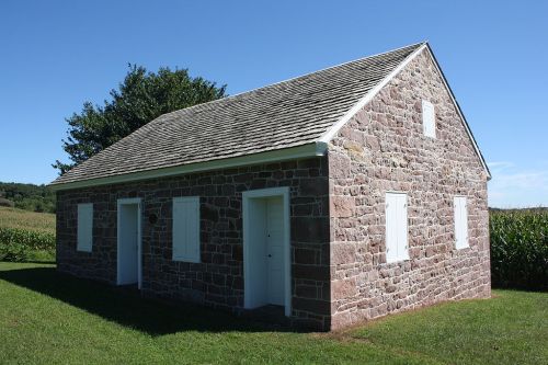 A stone house with white shutters, set against a clear blue sky and green grass, surrounded by fields.