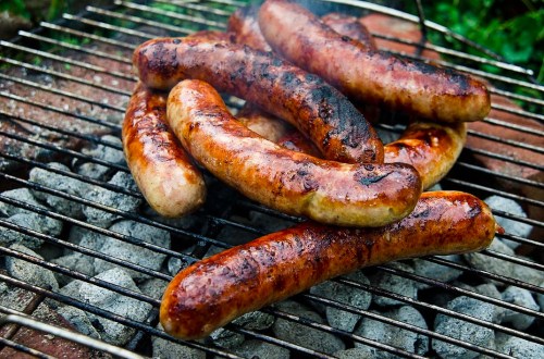 Grilled sausages sizzling on a barbecue grill over hot coals, with smoke rising in the background.