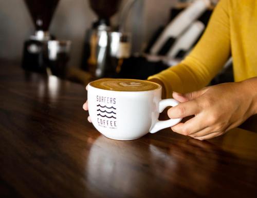 A person in a yellow sweater holds a large coffee cup with "Surfers Coffee" logo on a wooden counter.