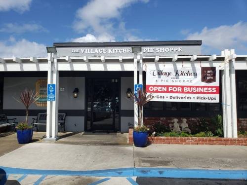 Exterior of Village Kitchen Pie Shoppe with a sign stating "Open for Business" and blue planters at the entrance.