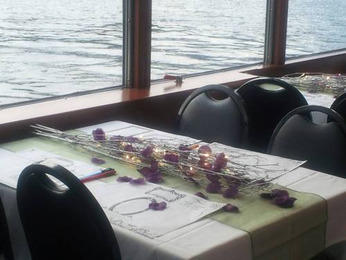 A table set with decorative branches and rose petals, overlooking a body of water through large windows.
