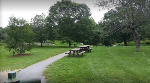 A peaceful park scene with picnic tables, green grass, and trees under a cloudy sky.