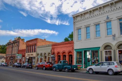 Historic storefronts line a street, featuring colorful buildings and parked cars under a blue sky with clouds.