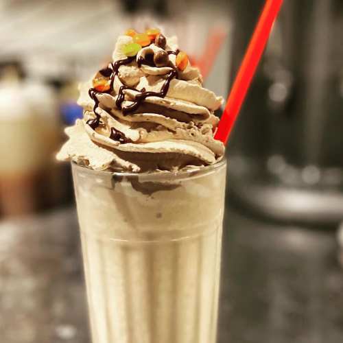A tall glass of chocolate milkshake topped with whipped cream, chocolate drizzle, and colorful candy pieces.