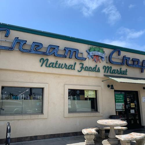 Exterior of Cream Crop Natural Foods Market, featuring a sign and outdoor seating area under a clear blue sky.