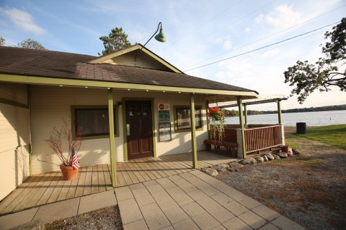 A cozy lakeside cabin with a wooden porch, potted plants, and a view of the water in the background.