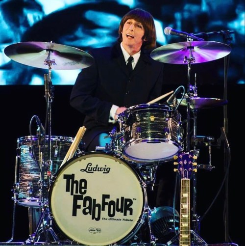 A drummer in a black suit performs on stage with a drum set labeled "The Fab Four" and a guitar nearby.