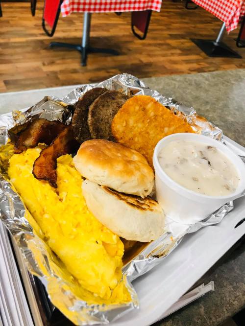A breakfast platter with scrambled eggs, bacon, sausage, hash browns, biscuits, and a side of gravy in a foil container.