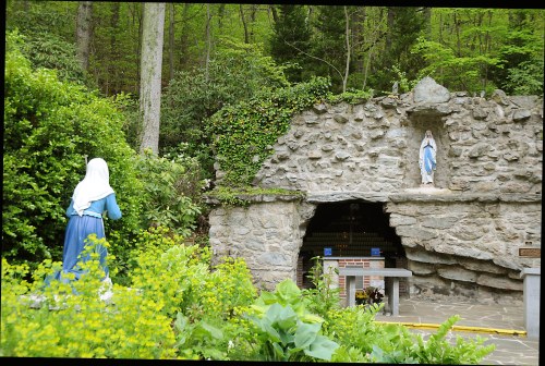 A stone grotto with a statue of Mary, surrounded by greenery and flowers, with a figure in a blue dress nearby.