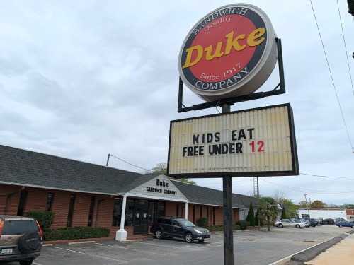 Sign for Duke Sandwich Company, stating "Kids eat free under 12," with a cloudy sky in the background.