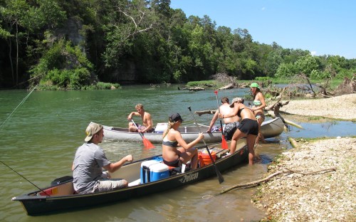 Two canoes on a river with people preparing to paddle, surrounded by lush greenery and a sunny sky.