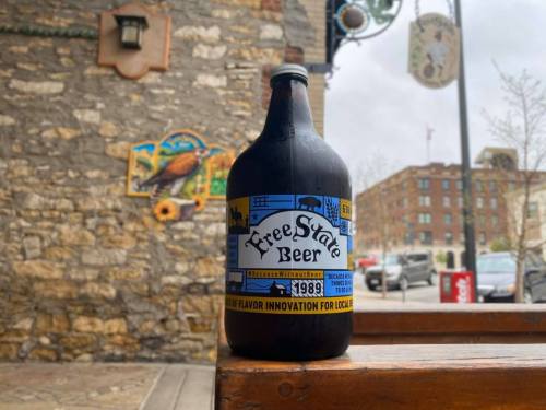 A black growler of FreeState Beer sits on a wooden table, with a stone wall and cityscape in the background.