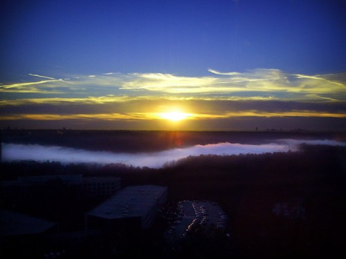 A vibrant sunset over a foggy landscape, with clouds illuminated in shades of orange and blue.