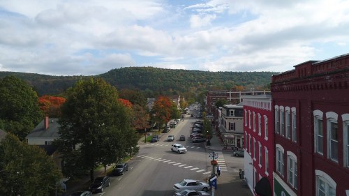 A scenic view of a small town street lined with trees, cars, and buildings, with a backdrop of colorful autumn foliage.