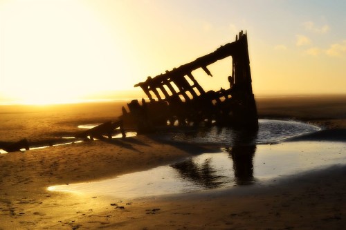 A shipwreck silhouette on a beach at sunset, partially submerged in shallow water, with soft golden light.