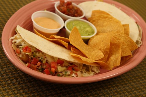 A pink plate with tacos, tortilla chips, and three dipping sauces: salsa, guacamole, and a creamy sauce.