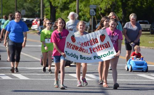Three girls hold a banner for the Georgia Strawberry Festival Teddy Bear Parade, with people walking behind them.