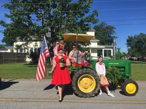 A group of people in colorful outfits poses on a green tractor, holding an American flag, during a sunny parade.