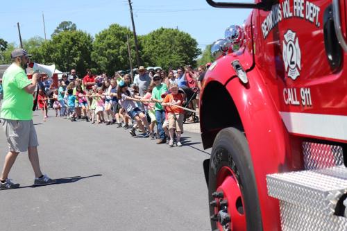 A crowd of children pulls a rope in a tug-of-war while a man with a megaphone encourages them near a fire truck.