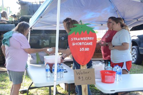 A booth selling strawberry shortcake and water at an outdoor event, with people interacting and a military vehicle in the background.