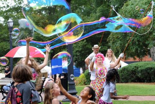 A colorful scene of children playing with bubbles created by a performer in a vibrant outfit at a park event.