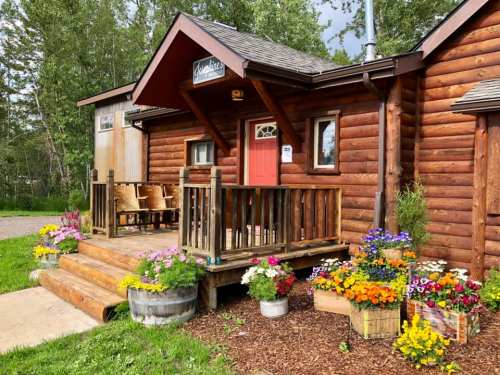 A cozy log cabin with a red door, surrounded by colorful flower beds and a wooden porch with chairs.