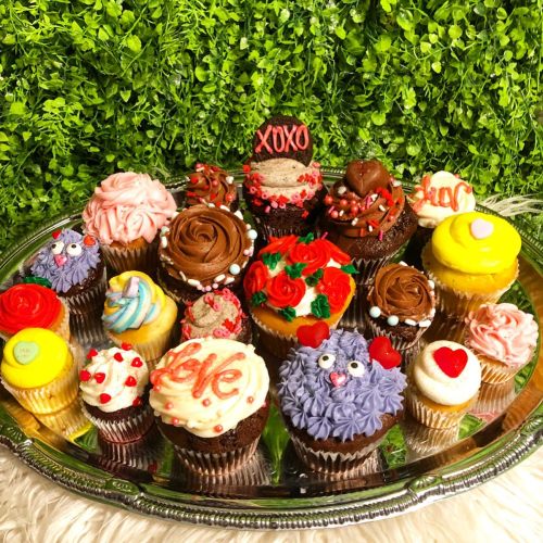 A variety of colorful cupcakes with intricate decorations, arranged on a silver tray against a green backdrop.
