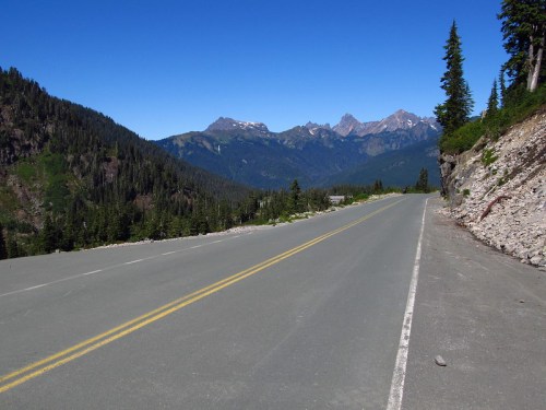 A winding road through a mountainous landscape, flanked by evergreen trees and clear blue skies.