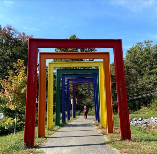 A person walks through a series of colorful, arch-shaped frames along a pathway in a scenic outdoor setting.
