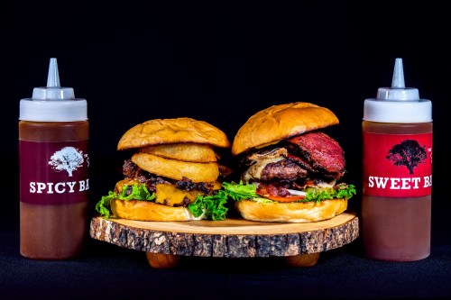 Two gourmet burgers on a wooden platter, one with spicy sauce and the other with sweet sauce, both topped with fresh ingredients.