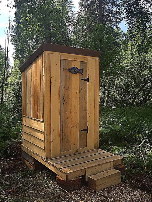 A wooden outdoor shed with a fish emblem, set in a lush green forest. Steps lead up to the door.