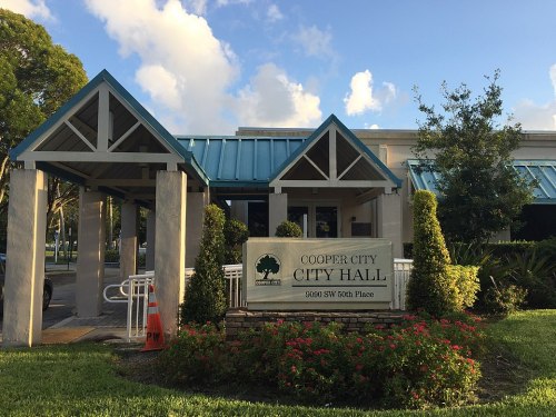 Cooper City Hall building with a blue roof, surrounded by greenery and flowers, under a clear sky.