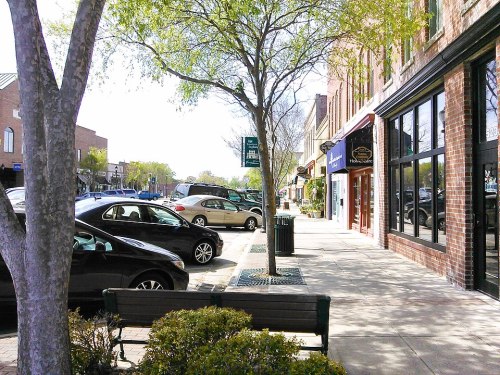 A sunny street scene with parked cars, trees, and shops along a sidewalk, inviting for a leisurely stroll.