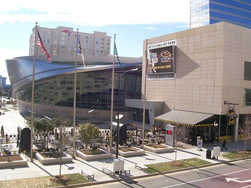 Exterior view of the NASCAR Hall of Fame, featuring flags and nearby restaurants, with a clear blue sky.