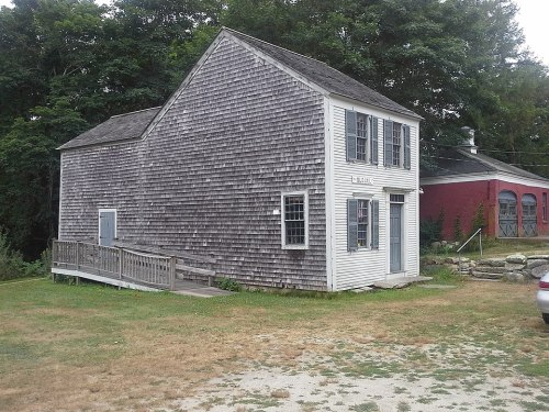 A weathered, two-story building with a shingled exterior and a ramp, surrounded by grass and trees.