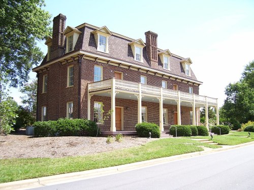 A large brick house with a porch, surrounded by greenery and trees, on a sunny day.