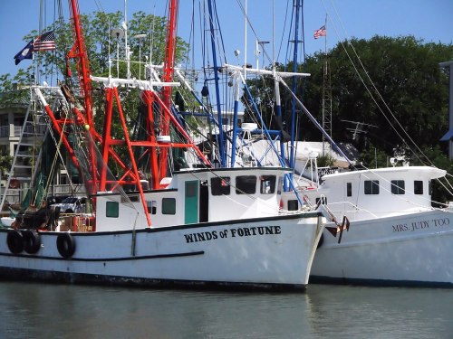 Two fishing boats docked in a harbor, one named "Winds of Fortune" and the other "Mrs. Judy Too," with masts and flags.