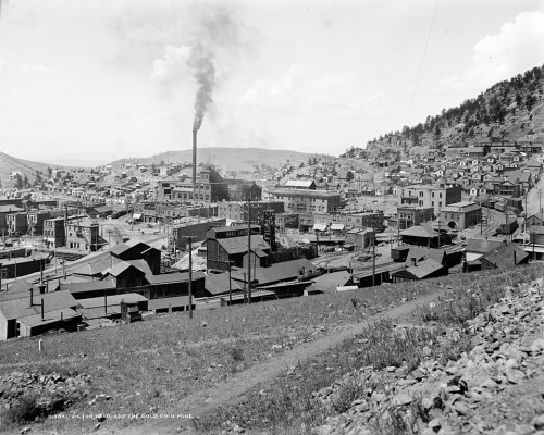 A historic black-and-white view of a town with buildings, smokestacks, and hills in the background.