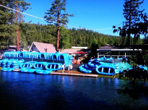 A sunny lakeside scene with blue rafts stacked on the shore, surrounded by trees and buildings.
