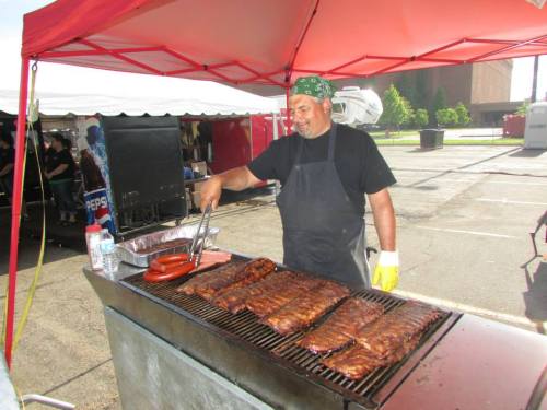 A man in a bandana and apron grills ribs and sausages at an outdoor event under a red tent.