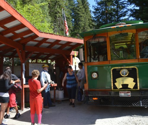 A green trolley bus parked at a rustic station, with people preparing to board in a forested area.