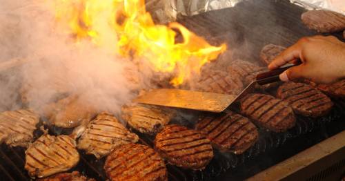 A hand flips sizzling burgers on a grill, with flames and smoke rising around them.