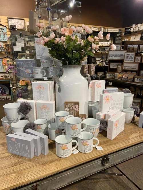 A display of decorative mugs, gift boxes, and a vase with pink flowers on a wooden table in a store.