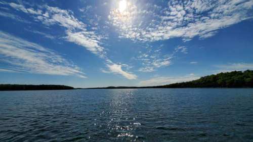 A serene lake under a bright sun, with scattered clouds and lush greenery along the shoreline.