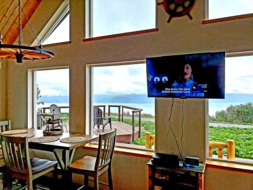 Cozy interior with a dining table, TV showing a program, and large windows overlooking a scenic outdoor view.