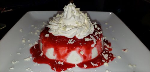 A creamy dessert topped with whipped cream and raspberry sauce, garnished with white chocolate shavings on a square plate.
